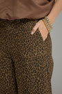 Close-up of a person wearing leopard-print pants with a brown top and stacked pearl bracelets, standing against a plain gray background.
