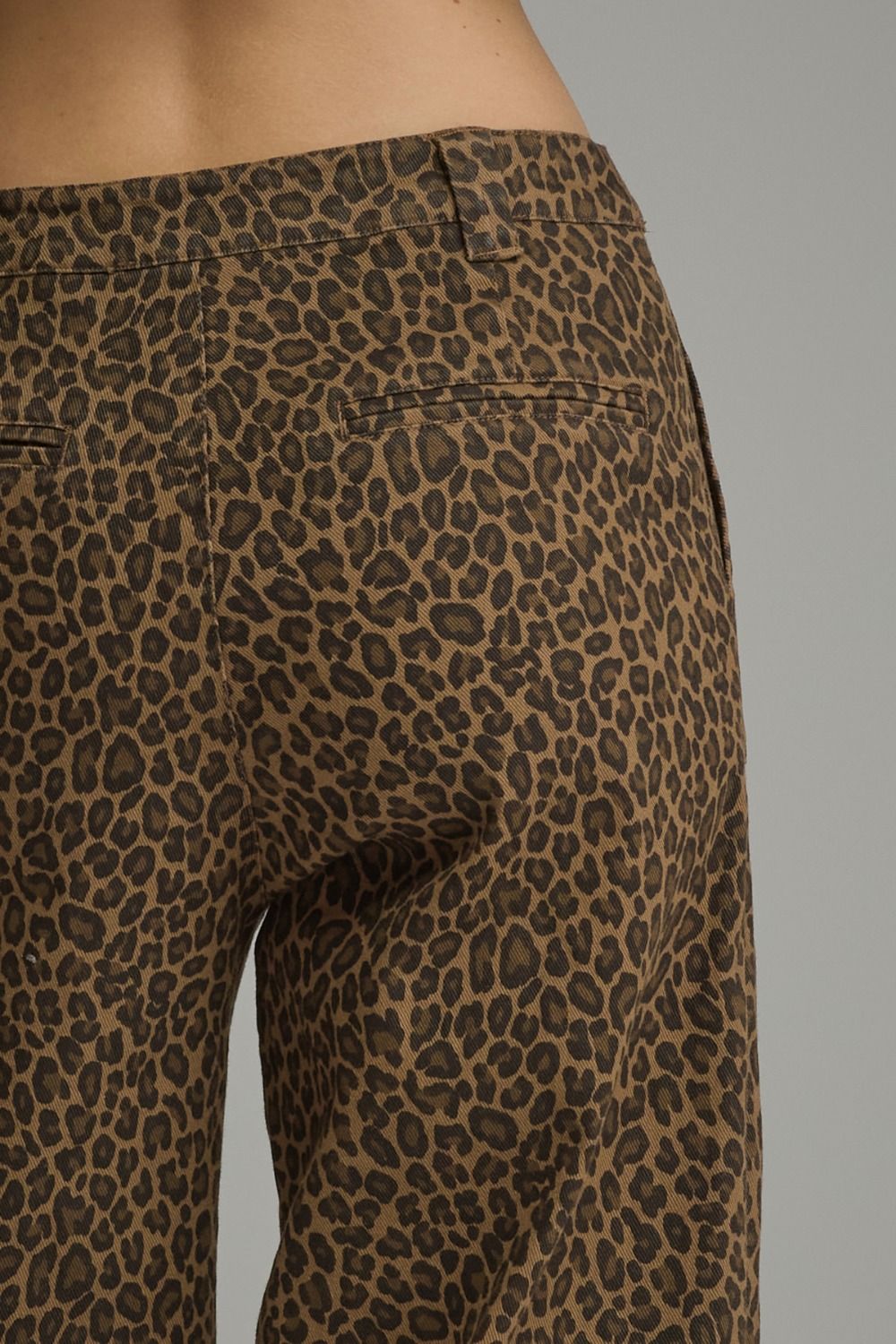 Close-up of a person wearing leopard-print pants, showing the back waistband, belt loops, and upper leg area against a plain gray studio background.