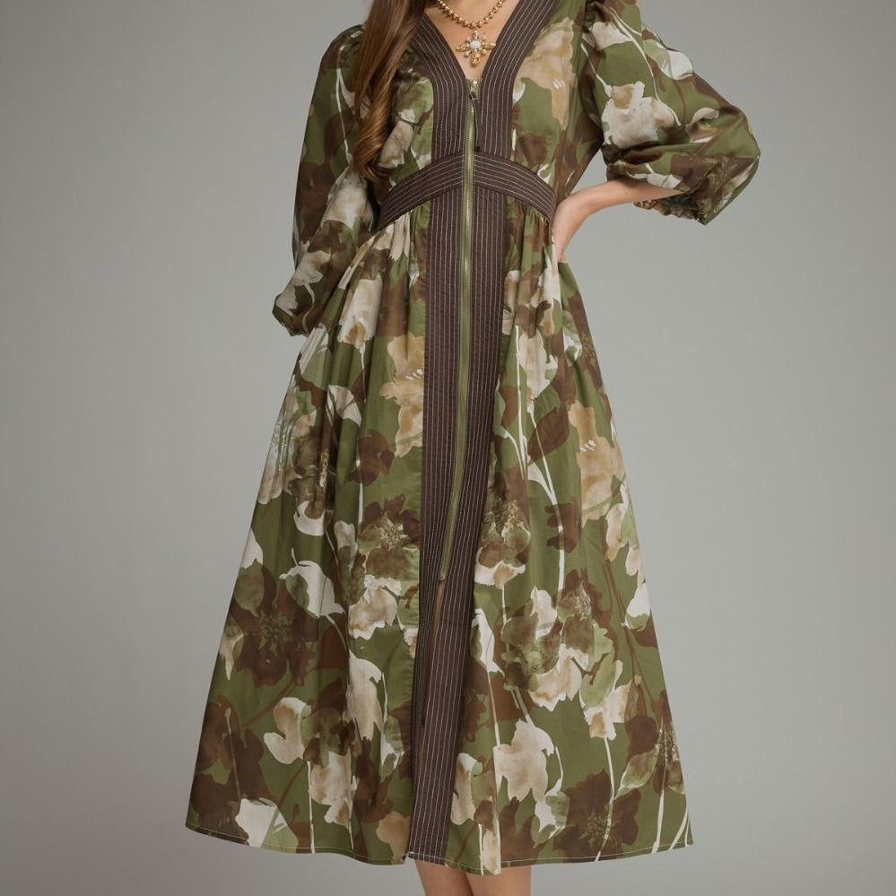 A woman models a long-sleeved midi dress with a green, brown, and beige floral print, featuring a dark brown vertical stripe down the front and a matching dark brown waist tie, set against a solid gray studio background.