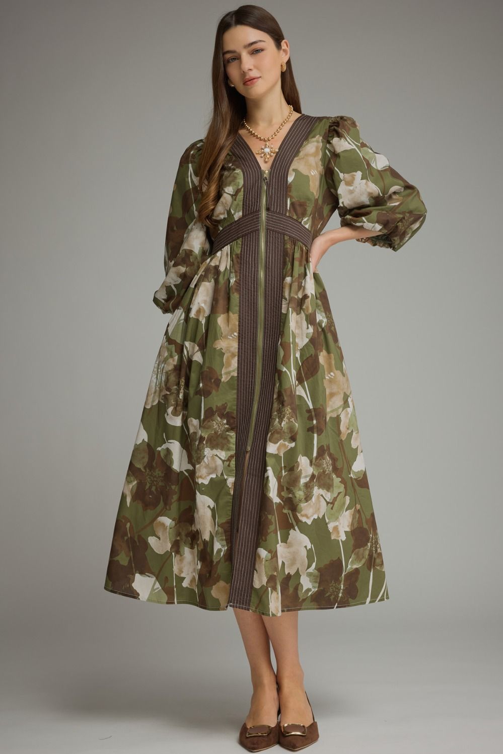 A woman models a long-sleeved midi dress with a green, brown, and beige floral print, featuring a dark brown vertical stripe down the front and a matching dark brown waist tie, set against a solid gray studio background.