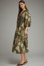 A woman modeling a midi-length floral print dress with a brown belt, styled with brown pointed-toe slingback heels against a neutral gray studio background.