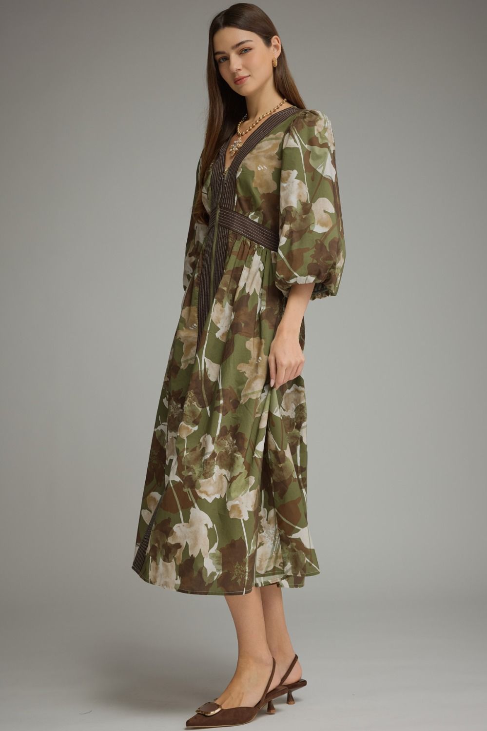 A woman modeling a midi-length floral print dress with a brown belt, styled with brown pointed-toe slingback heels against a neutral gray studio background.