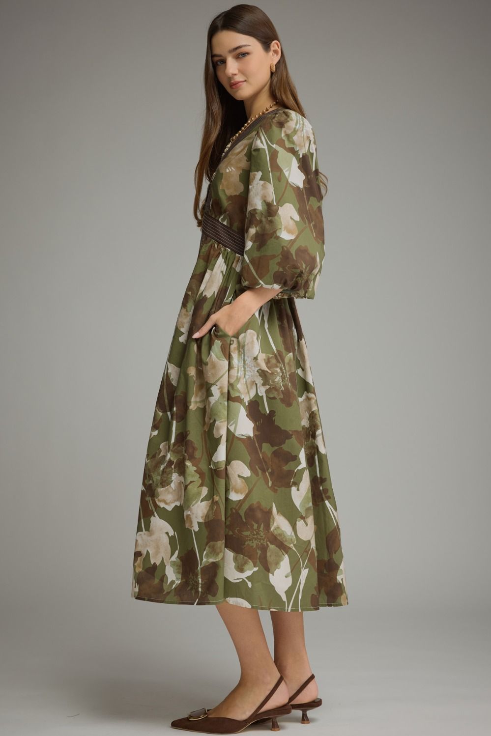 A woman modeling a knee-length olive green floral dress with a dark brown belt, standing against a neutral gray studio background.