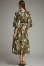 A woman viewed from the back wearing a knee-length dress with a green, brown, and beige floral camouflage pattern and a thin dark waist belt, standing against a plain gray studio background.