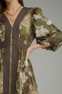 A close-up of a woman wearing a long-sleeved camouflage dress with dark brown vertical pleated trim down the front and along the waist, paired with a gold necklace.