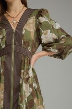 A close-up of a woman wearing a long-sleeved camouflage dress with dark brown vertical pleated trim down the front and along the waist, paired with a gold necklace.