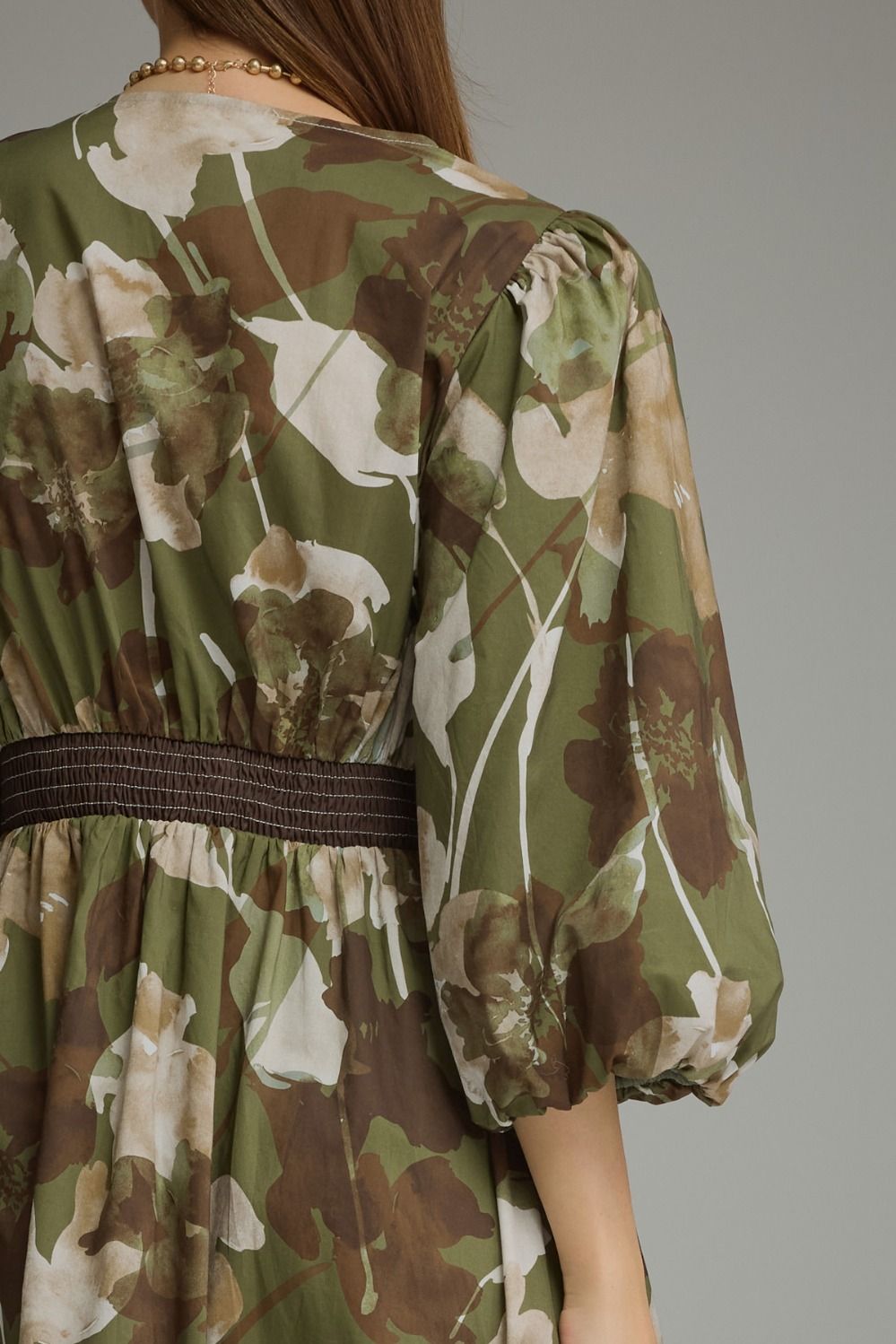 A close-up of a woman wearing a green floral dress with brown and beige tones, featuring a smocked waist and puffed elbow-length sleeves, shown from the back against a neutral gray background.