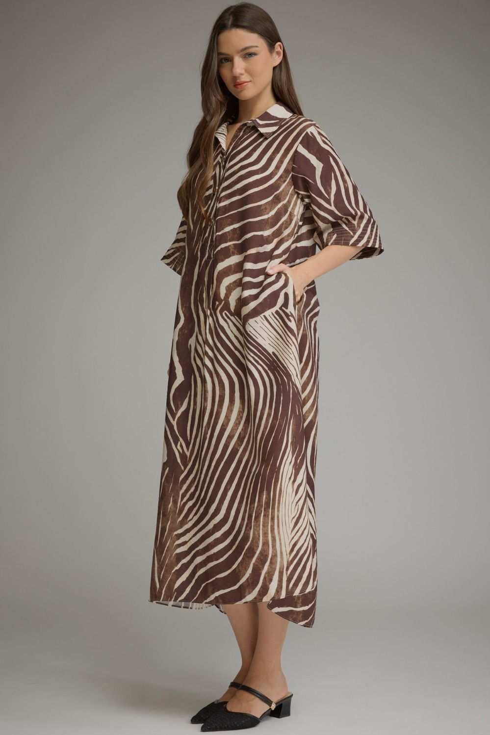 A woman modeling a brown and beige zebra-print midi shirt dress with three-quarter sleeves and a collar, paired with black flat shoes, against a neutral gray studio background.