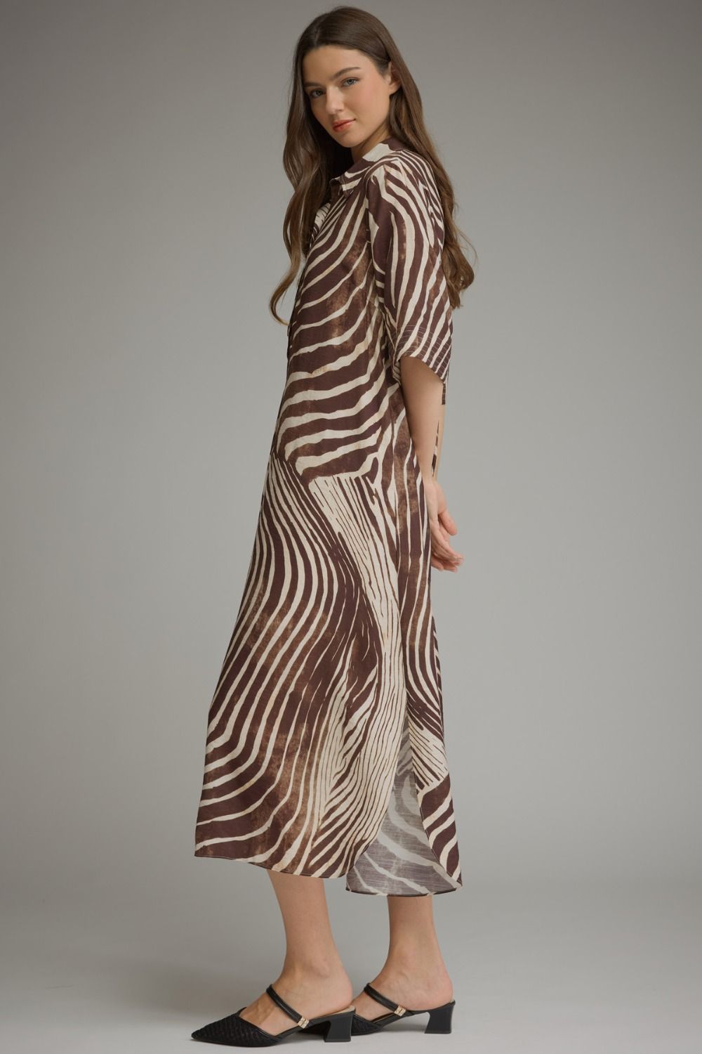 A woman modeling a brown and beige zebra-print midi dress with three-quarter sleeves, paired with black mule sandals, against a solid gray studio background.