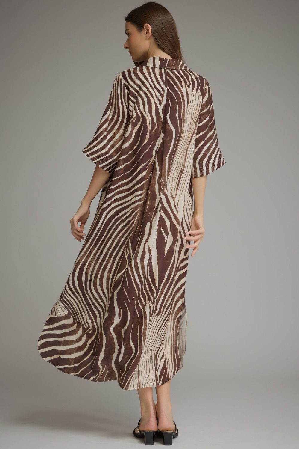 A woman modeling a brown and beige zebra-print kaftan dress, shown from the back against a neutral gray studio background.