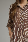 A close-up of a woman wearing a brown and cream zebra-print shirt dress with three-quarter sleeves and brown buttons, shown against a neutral gray background.