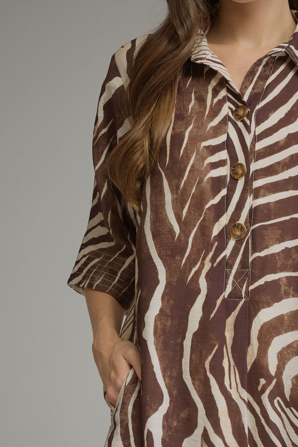 A close-up of a woman wearing a brown and cream zebra-print shirt dress with three-quarter sleeves and brown buttons, shown against a neutral gray background.