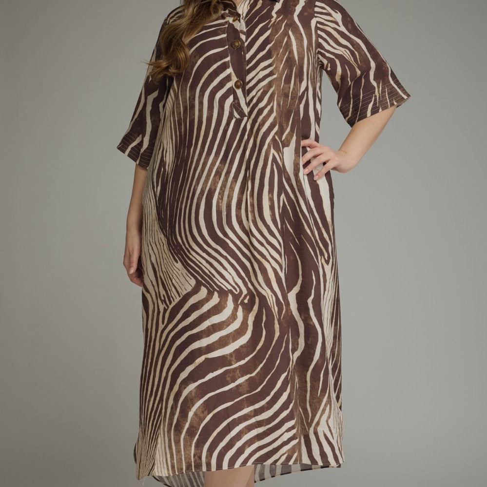 A woman modeling a brown and beige zebra-print midi shirt dress with short sleeves, a button-up front, and a relaxed fit, photographed against a neutral gray studio background.