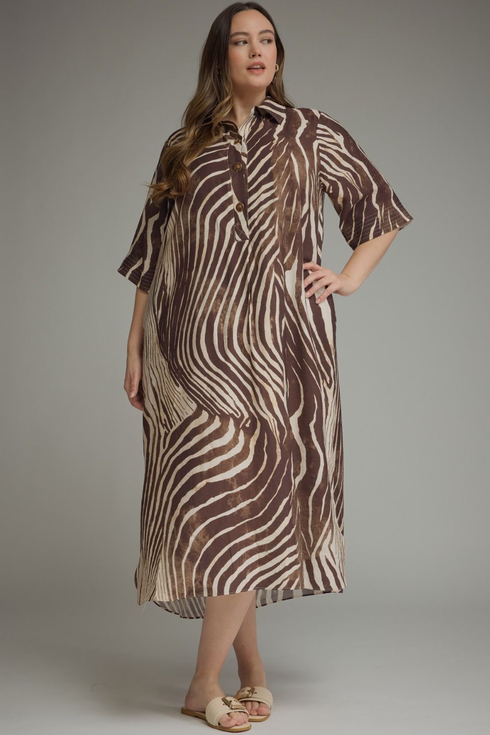 A woman modeling a brown and beige zebra-print midi shirt dress with short sleeves, a button-up front, and a relaxed fit, photographed against a neutral gray studio background.