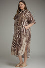 A woman modeling a brown and beige zebra-print midi dress with a high slit and short sleeves, paired with flat slide sandals, against a neutral gray studio background.