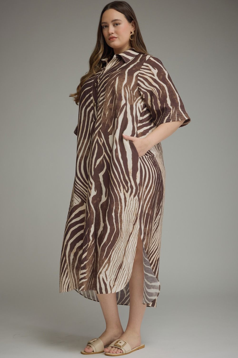 A woman modeling a brown and beige zebra-print midi dress with a high slit and short sleeves, paired with flat slide sandals, against a neutral gray studio background.