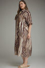 A woman modeling a brown and beige zebra-print midi dress with short sleeves and a high collar, paired with beige slide sandals, against a neutral gray studio background.