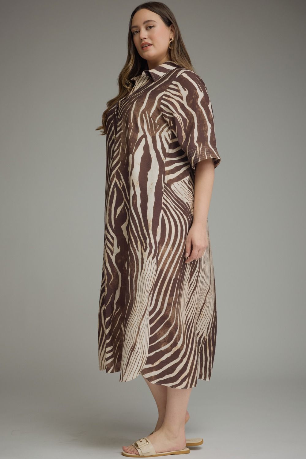 A woman modeling a brown and beige zebra-print midi dress with short sleeves and a high collar, paired with beige slide sandals, against a neutral gray studio background.