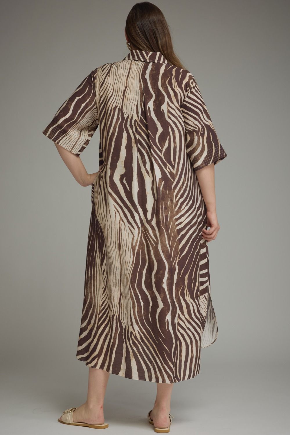 A woman viewed from the back wearing a mid-length brown and beige zebra-print shirt dress, paired with tan slide sandals, against a neutral gray studio background.