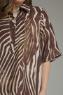 A close-up of a woman wearing a brown and cream zebra-print short-sleeve button-up shirt. The shirt features a textured, slightly mottled print with dark brown and warm cream stripes, accented by dark brown buttons.