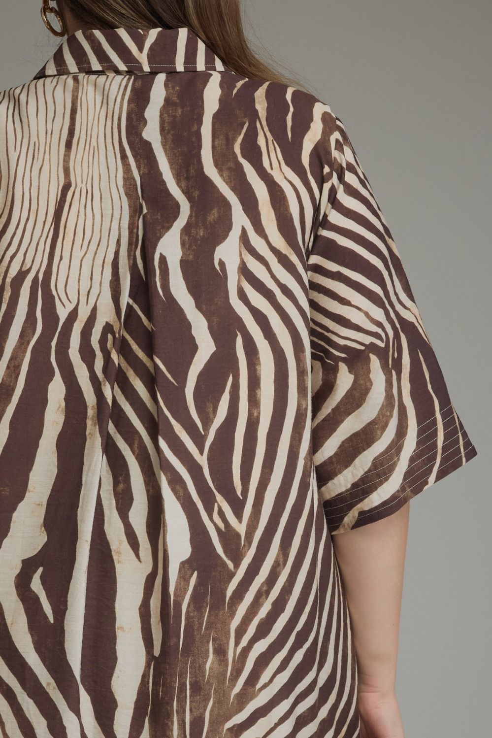 A close-up of a person wearing a short-sleeve button-up shirt with a brown and beige zebra-like abstract pattern, shown from the back against a plain gray background.