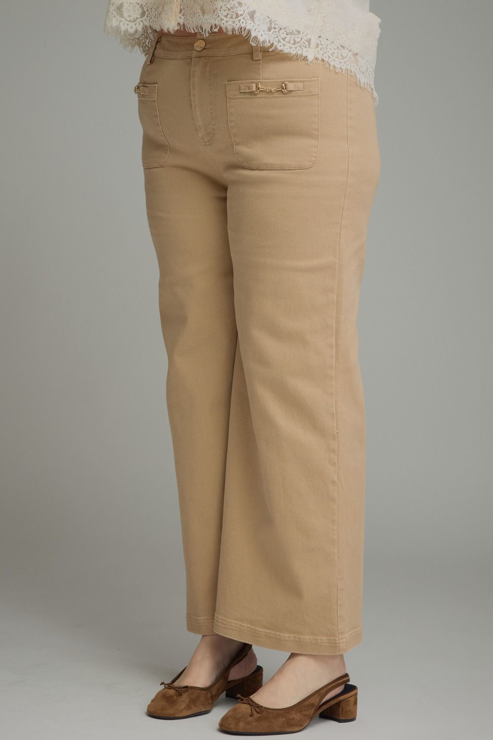 A person wearing tan cropped wide-leg pants with decorative front button details, paired with brown mule heels and a white lace-trim top, photographed against a plain light gray studio background.