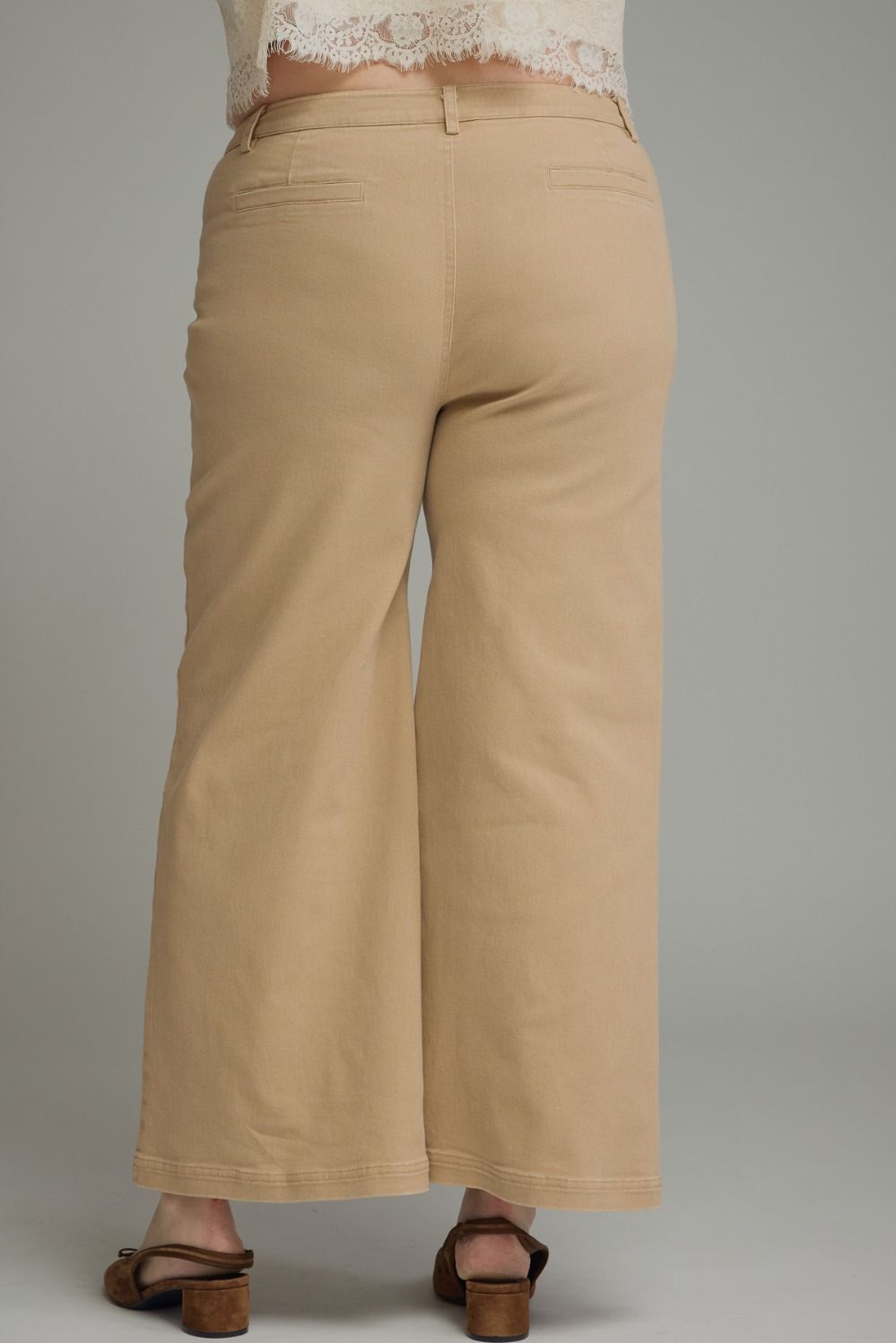 A person shown from the back wearing beige wide-leg cropped pants, a white lace-trim top, and brown heeled sandals against a plain gray studio background.