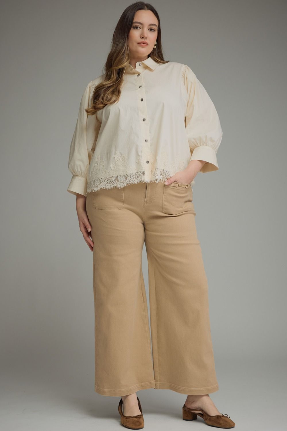A woman modeling a cream-colored blouse with lace trim and beige wide-leg pants against a solid gray studio background.