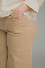 Close-up of a person wearing beige trousers with a decorative gold-tone horsebit detail on the front pocket, paired with a cream lace-trim blouse against a plain gray background.