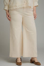 A person wearing cream wide-leg cropped pants with a matching cream lace-trim blouse and brown mule heels, photographed against a plain light gray studio background.