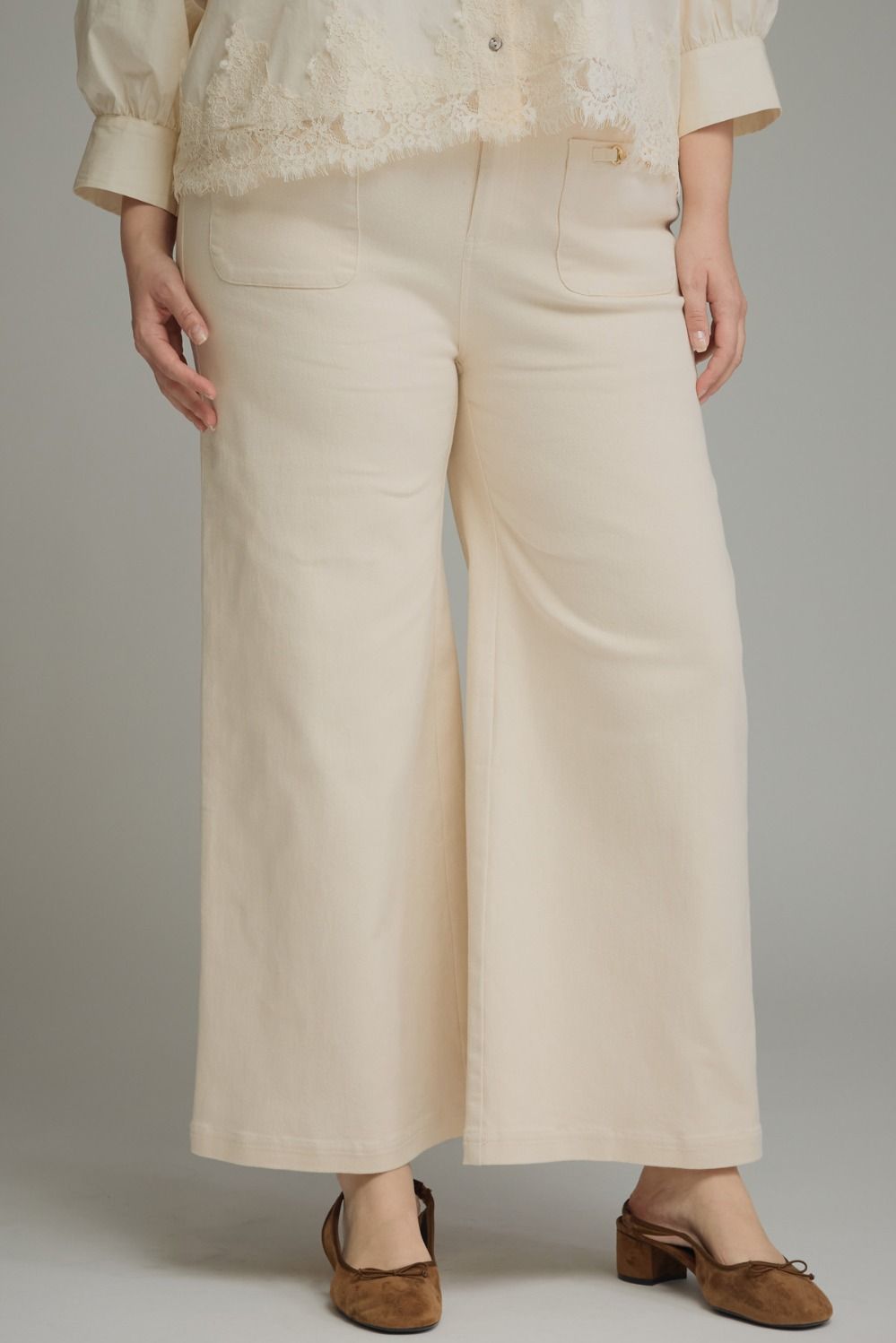 A person wearing cream wide-leg cropped pants with a matching cream lace-trim blouse and brown mule heels, photographed against a plain light gray studio background.