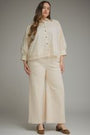 A woman modeling a cream-colored two-piece outfit consisting of a button-up blouse with lace trim and matching wide-leg cropped pants, paired with brown flat shoes, against a neutral gray studio background.
