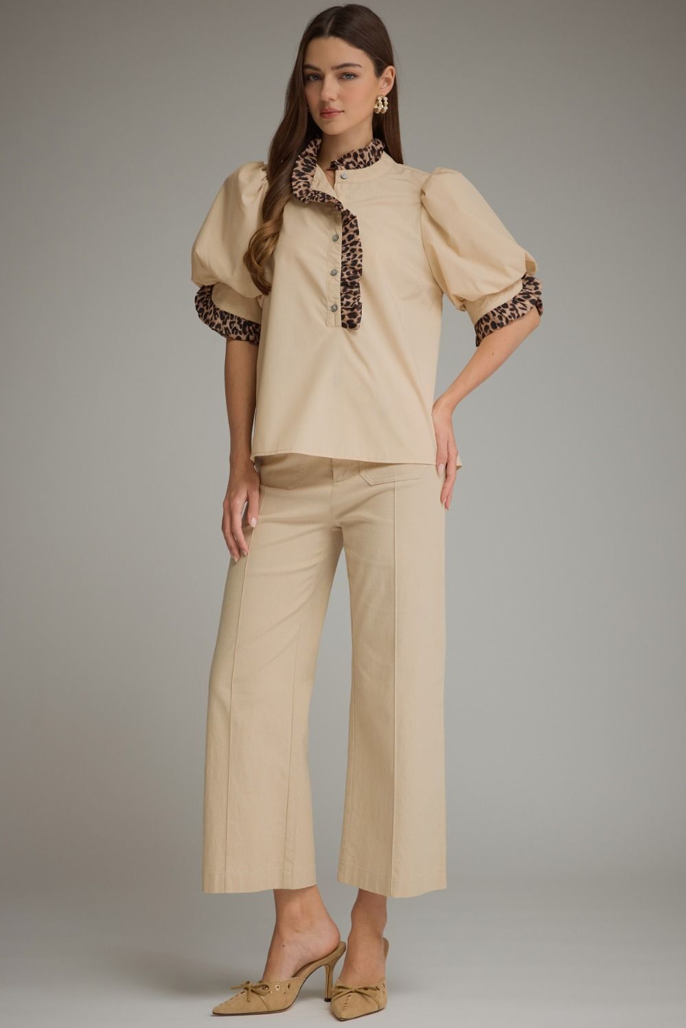 A woman models a beige two-piece set consisting of a short-sleeve blouse with leopard-print trim on the collar and cuffs, paired with matching beige wide-leg cropped pants and beige pointed-toe heels, against a solid gray studio background.