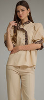 A woman modeling a beige blouse with leopard-print trim on the collar, placket, and puff sleeves, paired with matching beige trousers, against a solid gray studio background.