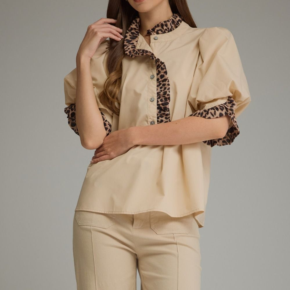 A woman modeling a beige blouse with leopard-print trim on the collar, placket, and puff sleeves, paired with matching beige trousers, against a solid gray studio background.
