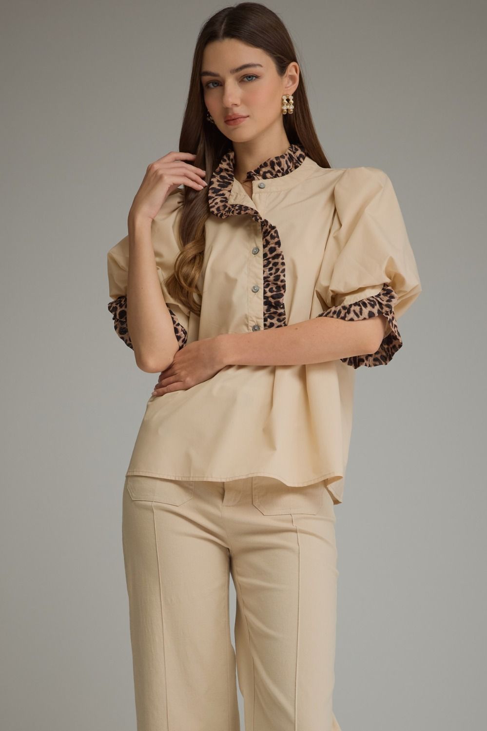 A woman modeling a beige blouse with leopard-print trim on the collar, placket, and puff sleeves, paired with matching beige trousers, against a solid gray studio background.