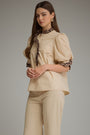 A woman modeling a beige puff-sleeve blouse with a leopard-print ruffled collar and cuffs, paired with matching beige trousers, against a solid gray studio background.