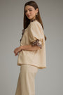 A woman modeling a beige short-sleeve blouse with leopard-print ruffle trim at the collar and sleeve cuffs, paired with matching beige trousers, against a plain gray studio background.
