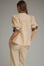 A woman shown from the back wearing a beige blouse with leopard-print ruffled trim at the collar and sleeve cuffs, paired with matching beige trousers, against a plain gray studio background.