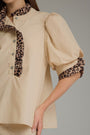 A close-up of a beige blouse with leopard-print ruffled trim on the collar and sleeve cuffs, shown on a model against a neutral gray background.