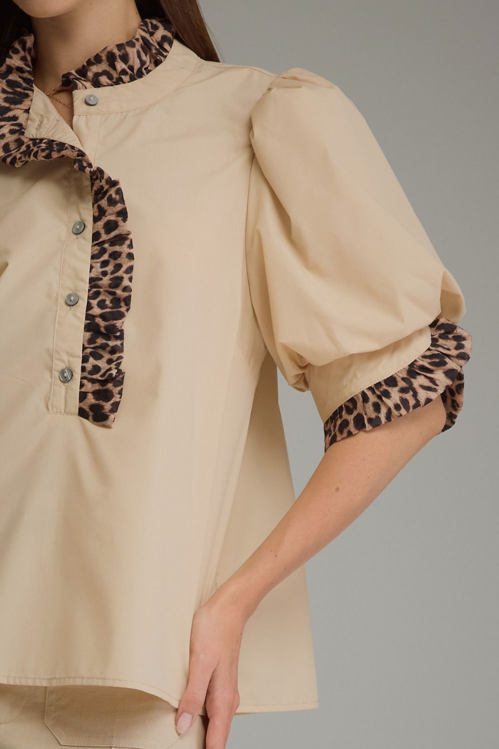 A close-up of a beige blouse with leopard-print ruffled trim on the collar and sleeve cuffs, shown on a model against a neutral gray background.