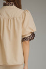 A close-up back view of a woman wearing a beige blouse with leopard-print trim on the collar and sleeve cuffs, set against a plain gray studio background.