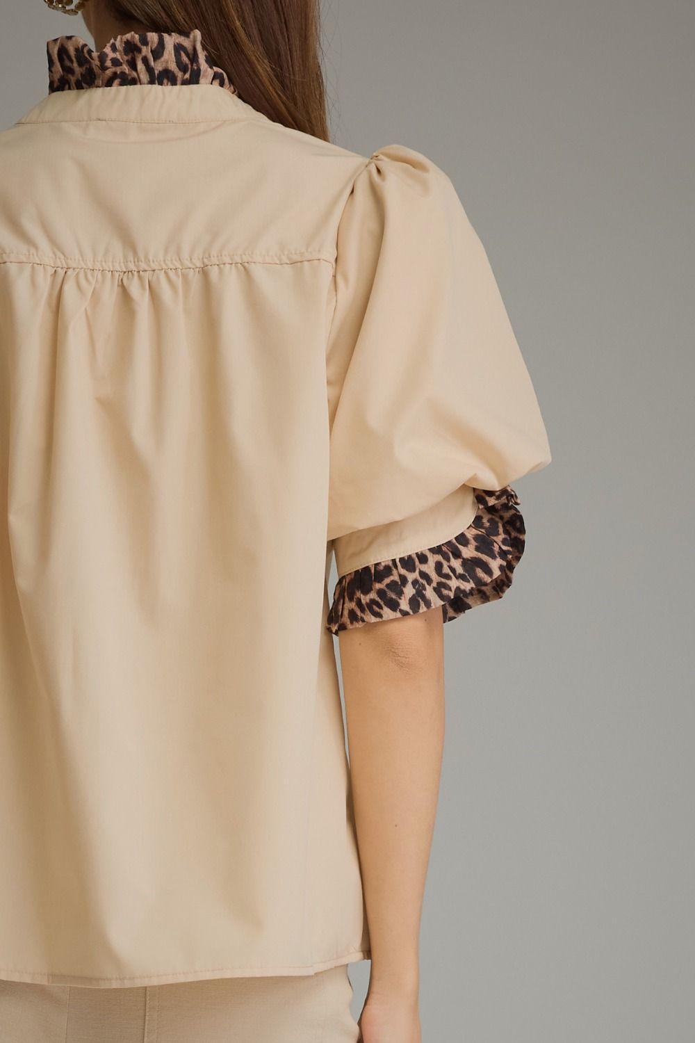 A close-up back view of a woman wearing a beige blouse with leopard-print trim on the collar and sleeve cuffs, set against a plain gray studio background.
