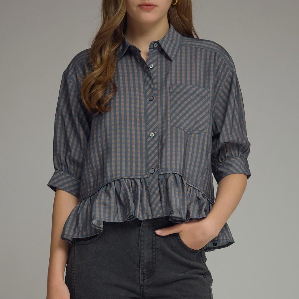 Woman wearing a navy checkered blouse with ruffled bottom and black pants on a gray background