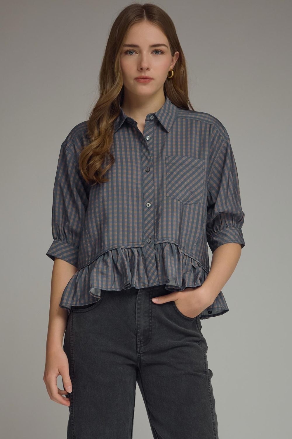 Woman wearing a navy checkered blouse with ruffled bottom and black pants on a gray background