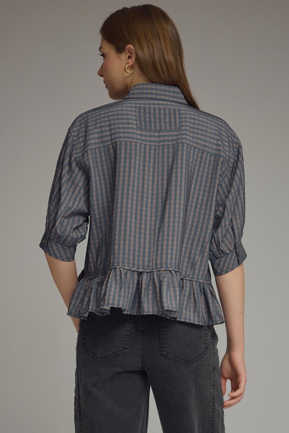 Woman wearing a gray checkered shirt with ruffled bottom and dark pants on a gray background