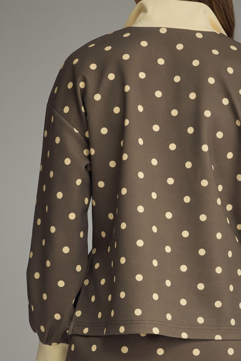 A close-up back view of a brown long-sleeve top with a cream polka dot pattern, shown on a mannequin against a neutral gray background.