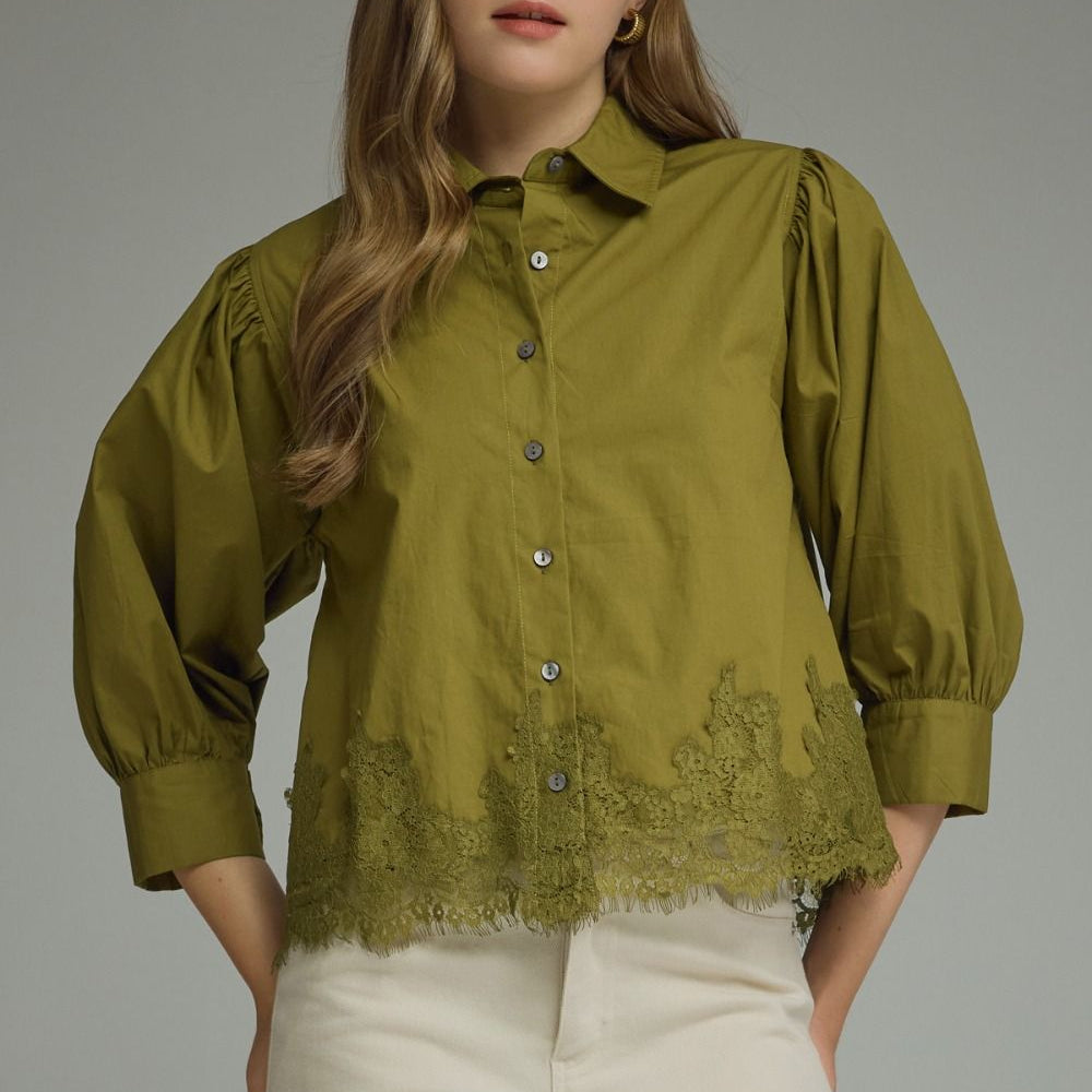 A woman wearing an olive green button-up blouse with puff sleeves and a lace hem, paired with cream-colored pants, against a solid gray studio background.