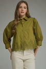 A woman wearing an olive green button-up blouse with puff sleeves and a lace hem, paired with cream-colored pants, against a solid gray studio background.