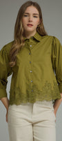 A woman wearing an olive green button-up blouse with puff sleeves and a lace hem, paired with cream-colored pants, against a solid gray studio background.